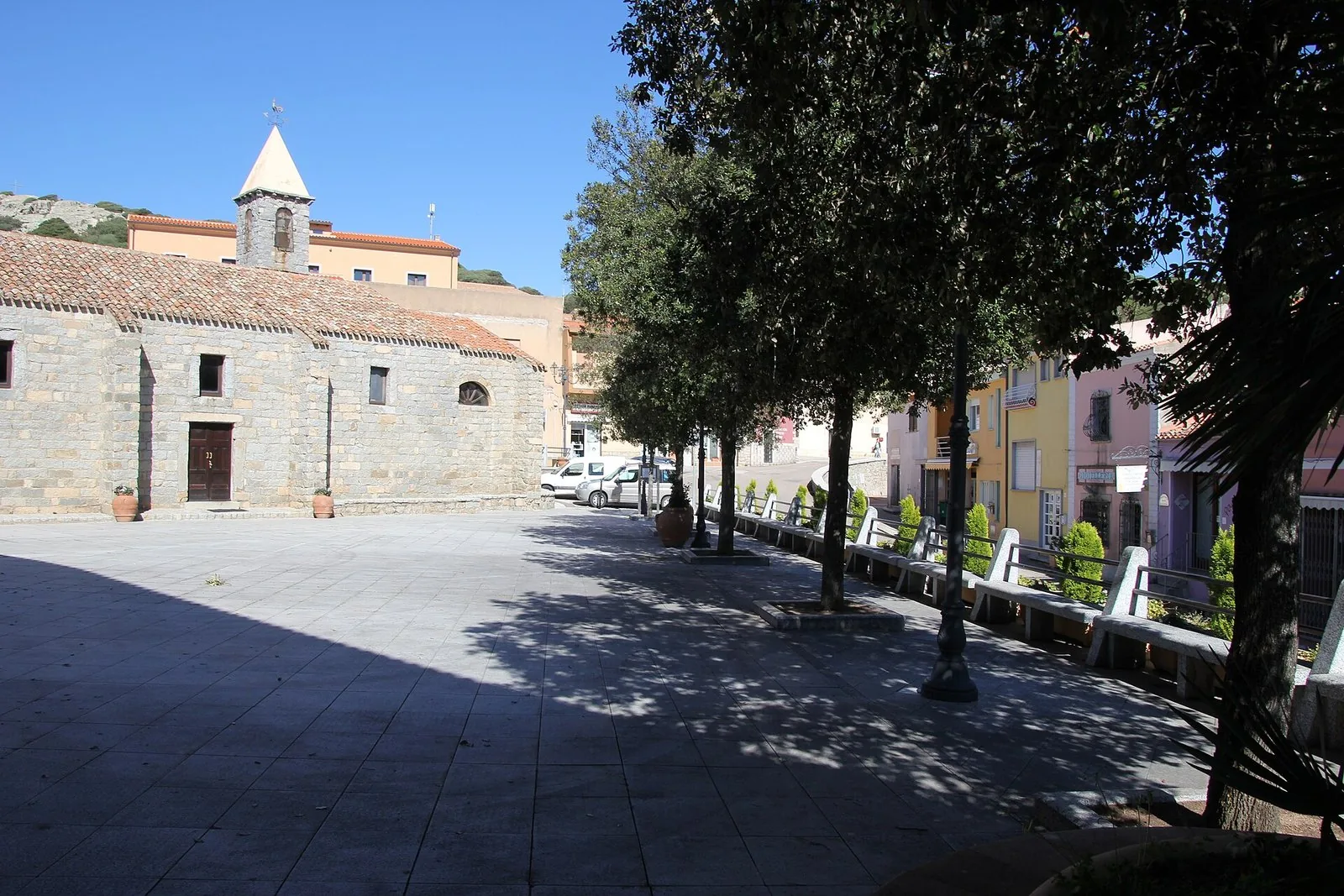 Piazza di Trinità d'Agultu con una chiesa in blocchi di granito e il campanile, un filare di alberi e case colorate sulla destra